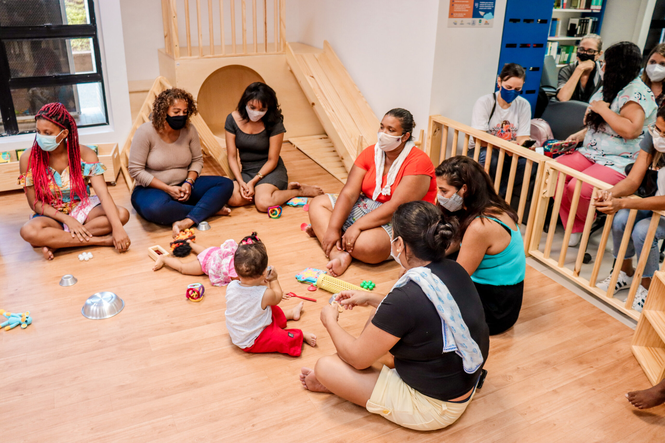Uma roda de mulheres está sentada num piso de madeira dentro de uma bebeteca. Alguns bebês estão sentados no chão brincando com objetos. Atrás, mobiliários pikler. 
